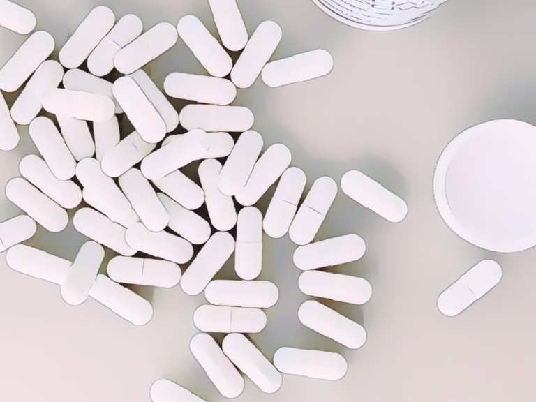A scattered pile of white oblong pills with a scored center, possibly supplements or medication, on a neutral surface. An open pill bottle and its white cap are partially visible in the background. Photo by The-Lore on Unsplash