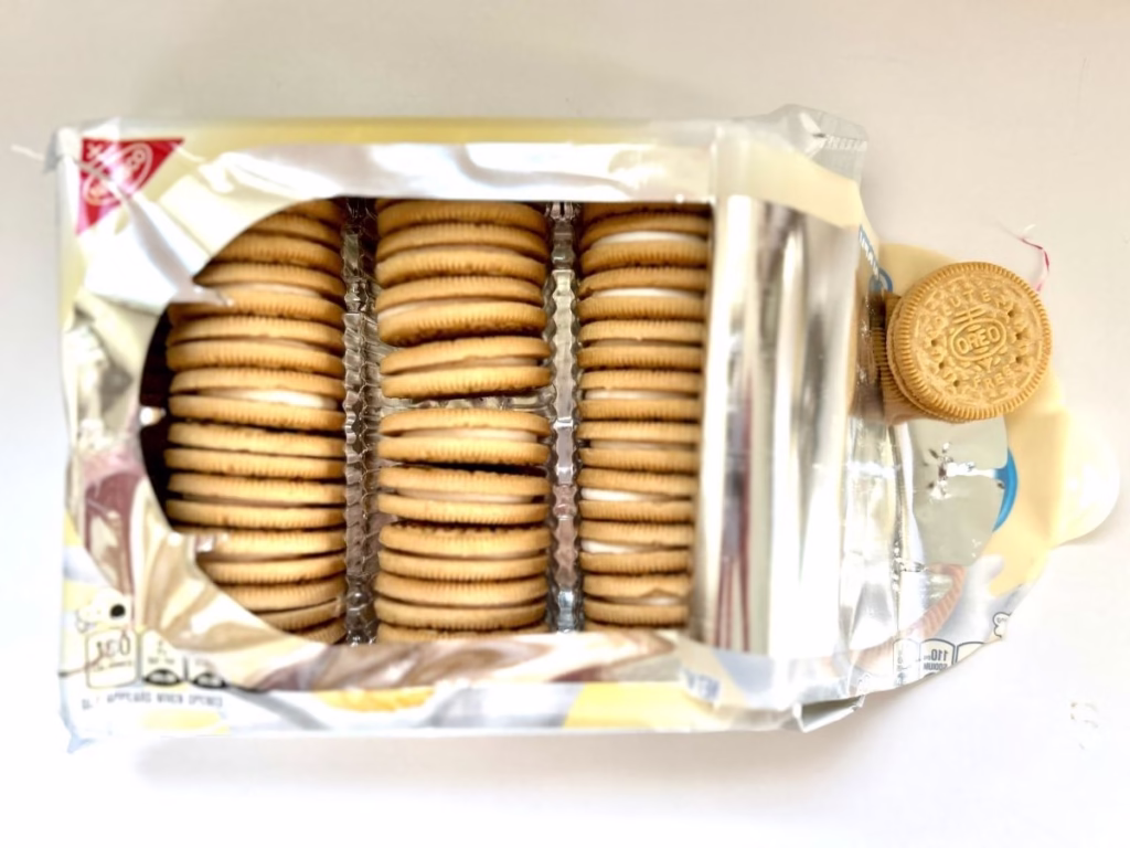 An opened package of "Golden Gluten Free OREO" sandwich cookies is shown from above. The silver foil-lined tray inside contains three rows of stacked vanilla sandwich cookies with white cream filling. One loose cookie is positioned to the right of the open package, next to some crumpled packaging. The white and yellow outer packaging is visible around the foil tray. Nutritional information can be seen on the bottom left corner of the packaging.