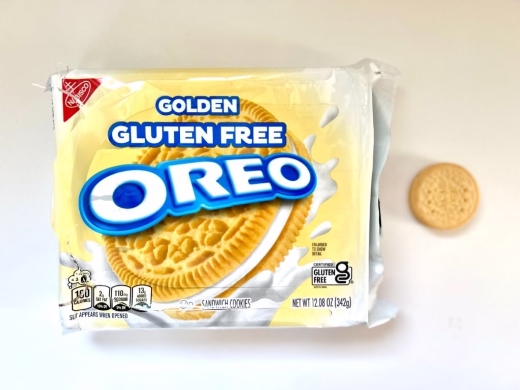A plastic package of "Golden Gluten Free OREO" sandwich cookies is displayed on a white surface. The packaging features a large image of the golden sandwich cookie with white cream filling, surrounded by splashes of milk. The "OREO" logo is prominently displayed in blue and white. On the right side of the package, a single Golden Oreo cookie is placed next to it. The packaging also shows nutritional information, including "150 Calories" per serving and "13g Added Sugars." A "CERTIFIED GLUTEN FREE" logo is visible. The net weight is stated as "NET WT 12.08 OZ (342g)." The Nabisco logo is in the top left corner.