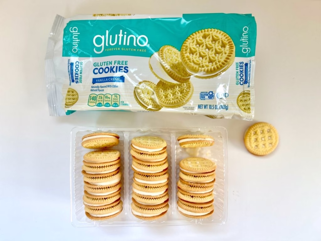 A package of "glutino FOREVER GLUTEN FREE COOKIES Vanilla Crème" is positioned at the top of the image. The packaging is light blue and white, featuring an illustration of golden sandwich cookies with cream filling. Below the sealed package, an opened clear plastic tray containing three stacks of glutino vanilla crème sandwich cookies is displayed. To the right of the tray, a single loose glutino cookie is placed. The background is a plain white surface.