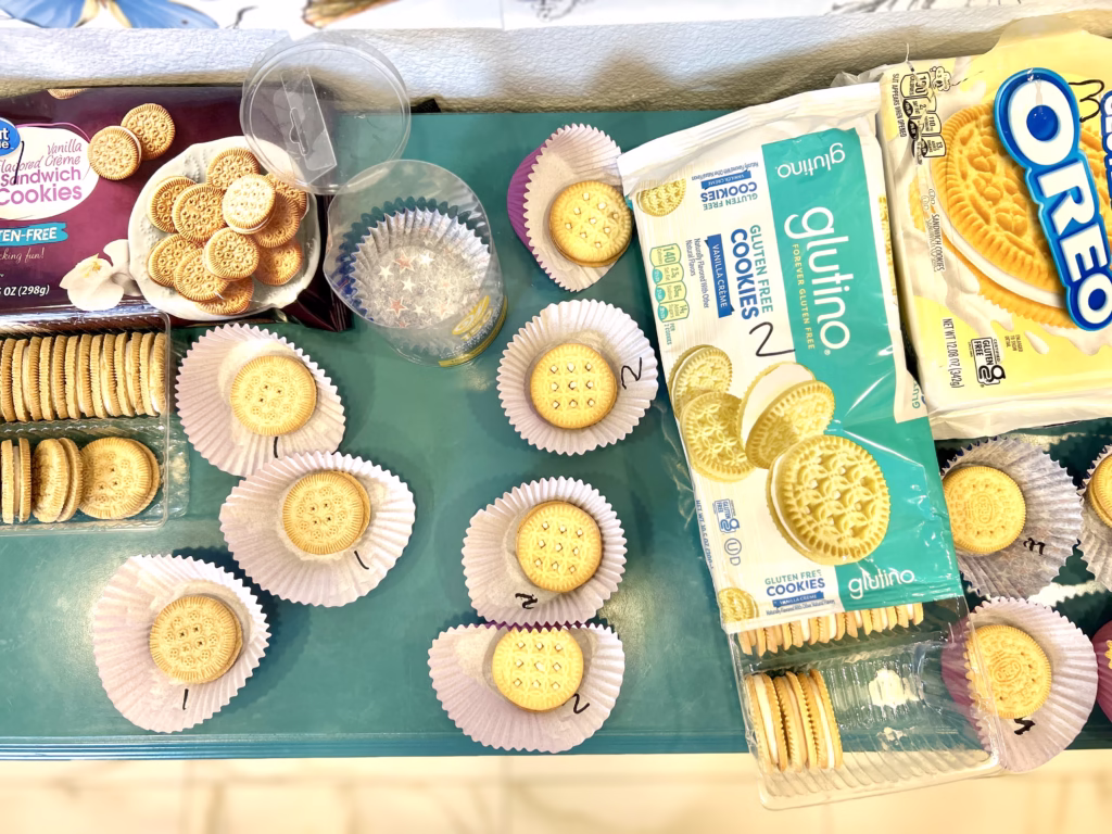 A flat lay shows gluten-free vanilla sandwich cookies and golden lemon Oreos being prepared for individual servings. The cookies are arranged in white and purple cupcake liners on a teal surface. On the left, a partially eaten box of "Great Value Gluten-Free Vanilla Sandwich Cookies" is visible, with a container holding several cookies. Below it, a plastic tray holds multiple rows of these vanilla sandwich cookies. In the center, numerous individual vanilla sandwich cookies are placed in white cupcake liners. Some empty clear plastic containers and a purple cupcake liner are also present. On the right, a large package of "Glutino Gluten-Free Vanilla Creme Cookies" is open, revealing more of the vanilla cookies. To its right, an open package of "OREO Golden Lemon Sandwich Cookies" shows the yellow cookies. Below this, several golden lemon Oreos are also individually placed in white and purple cupcake liners.